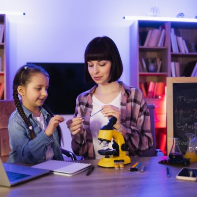 Woman sitting with girl at table and using microscope for studying sciences.